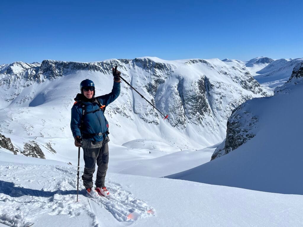 Ski Guide Rasmus Normann Levin Larsen står på toppen av et fjell på ski med neven i været og et smil om munnen. Det er sol og blå himmel og en alpin dal i bakgrunnen. 