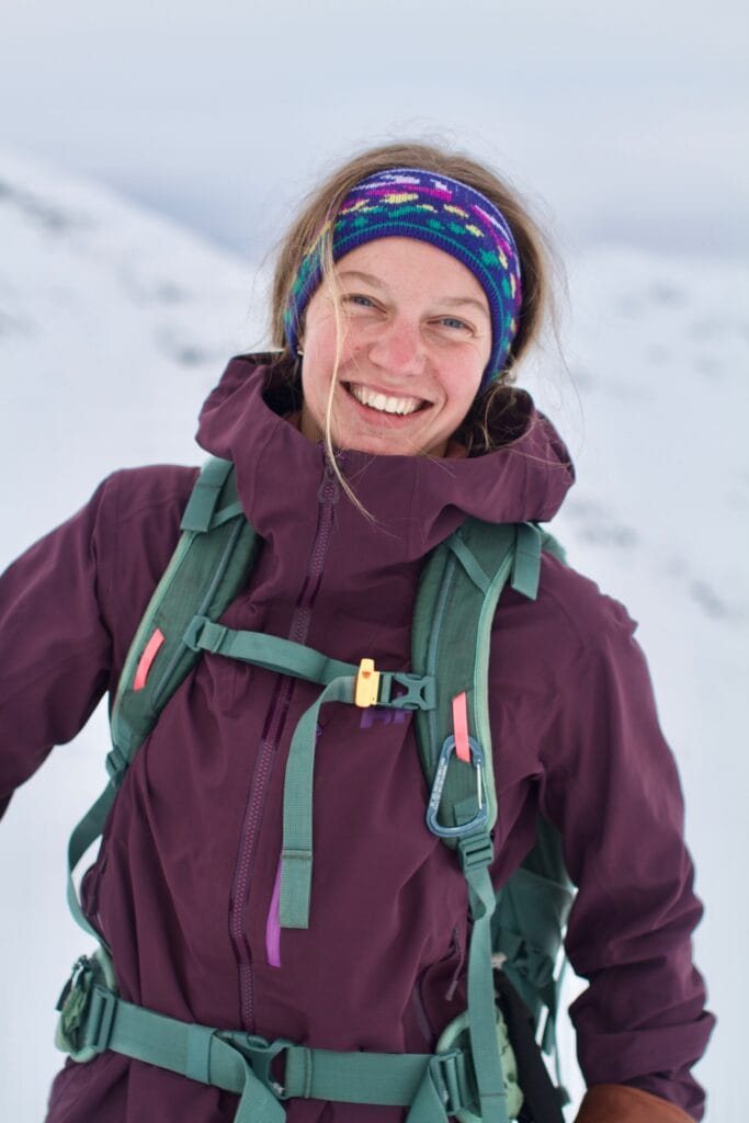 Guide Tabitha smiler ute på topptur med hvite fjell i bakgrunnen.