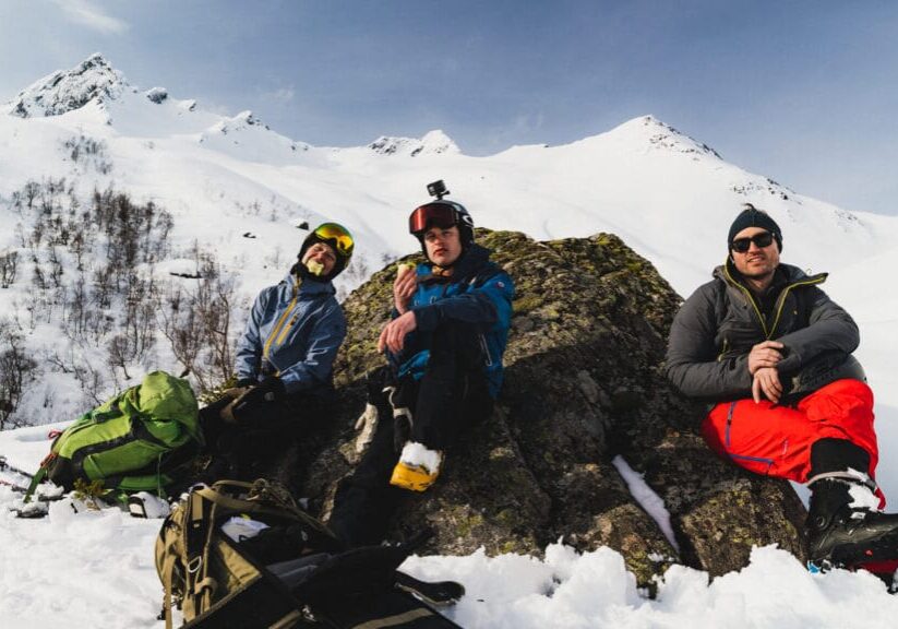 Guiden Tabitha med to venner har matpause mitt i vinterfjellet med sol i fleisen.