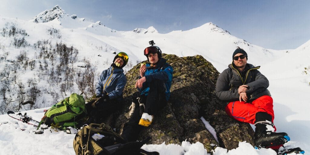 Guiden Tabitha med to venner har matpause mitt i vinterfjellet med sol i fleisen.